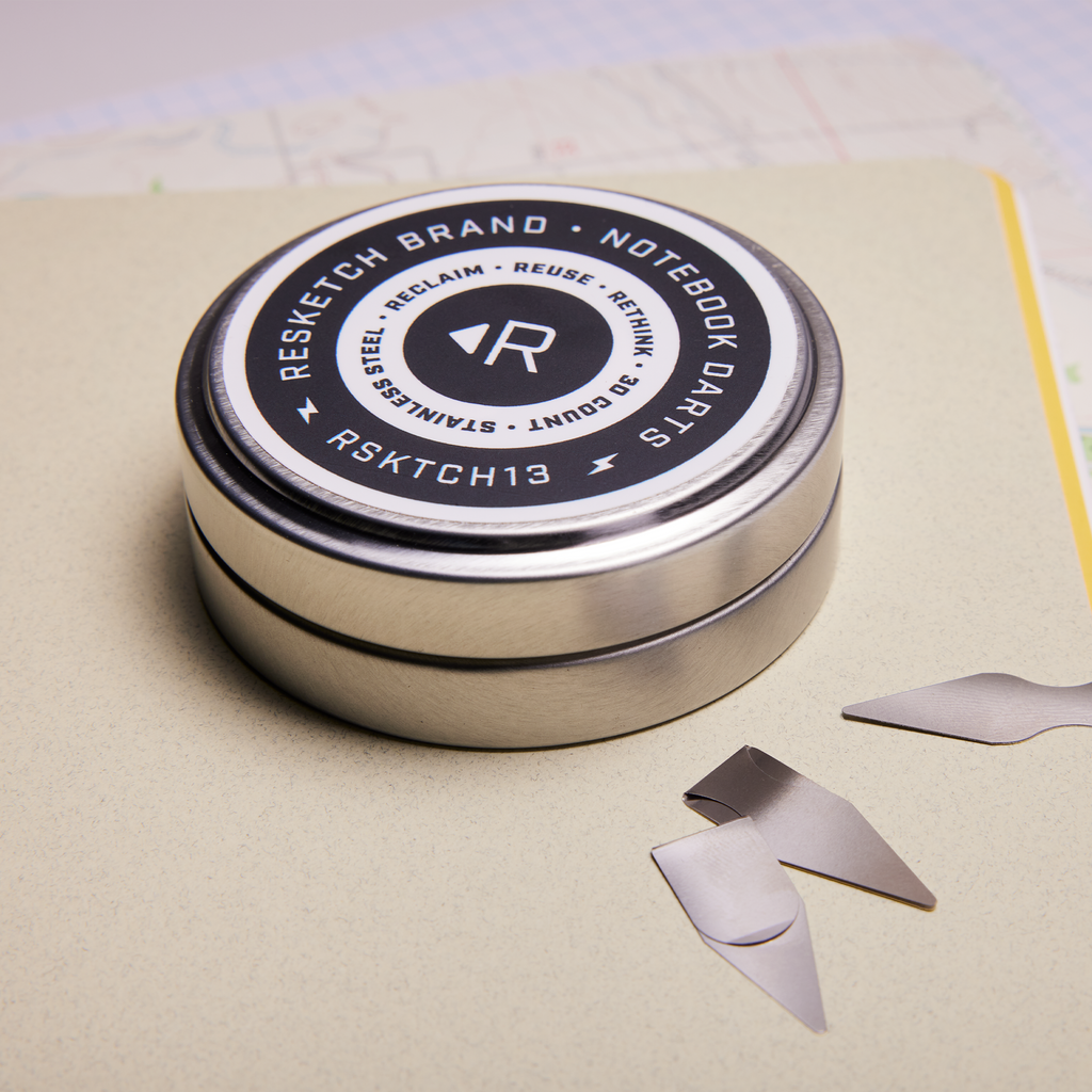 One can of Resketch notebook Darts sitting on a Resketch notebook, open to a a lined page, an map page and constrction paper page with three Stainless Steel darts 