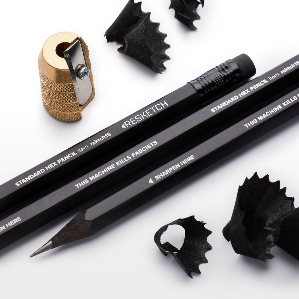 Brass pecil sharpener, three black resketch pencils on their side. Pencils are made from black wood, black eraser and a black Ferrule. Black pencil shavings The Machine Kills Fasicists printed on the side.