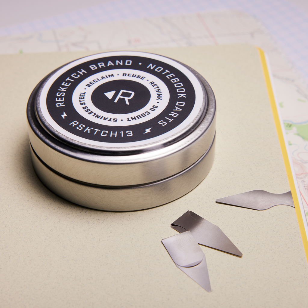 Lifestyle Picture of an Aluminum tin of Resketch Notebook Darts, sitting on top of a Resketch notebook made from 100% reclaimed, rescued and upcycled paper.